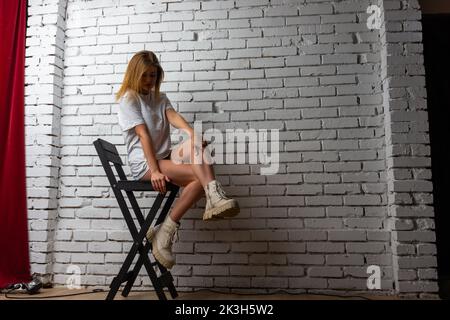 Portrait modische junge Frau trägt stilvolle Kleidung, weißes T-Shirt, schwarzen Rock, posiert auf Stuhl. Frau fesselte ihre Haare und posierte auf der Kamera Stockfoto