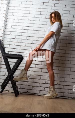 Portrait modische junge Frau trägt stilvolle Kleidung, weißes T-Shirt, schwarzen Rock, posiert auf Stuhl. Frau fesselte ihre Haare und posierte auf der Kamera Stockfoto