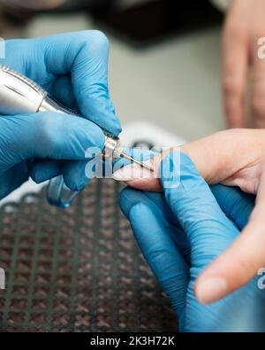 Verarbeiten Sie professionelle Hardware Maniküre. Master Manikurist in blauen Handschuhen arbeitet mit Fräser und entfernt alte Gelpolitur von weiblichen Client Nägel Stockfoto