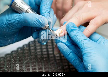 Verarbeiten Sie professionelle Hardware Maniküre. Manikuristin in blauen Handschuhen arbeitet mit Fräser und entfernt alte Gelpolitur von weiblichen Client-Nägeln. Wählen Stockfoto
