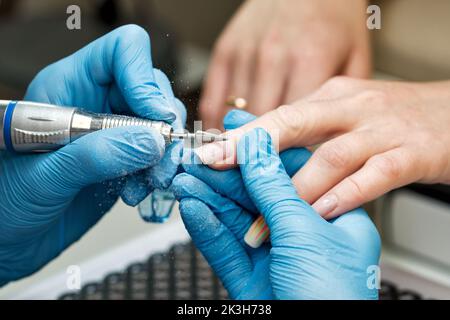 Prozess Hardware Maniküre. Manikuristin in blauen Handschuhen arbeitet mit Fräser und entfernt alte Gelpolitur von weiblichen Client-Nägeln. Professioneller Mani Stockfoto