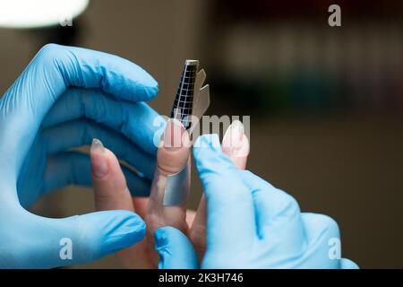 Konstruktion von Schönheitsnägeln. Untere Nagelverlängerung. Meister Manikurist im Schönheitssalon prüft, ob Papier Formen für Nagelverlängerung gleichmäßig fem installiert Stockfoto