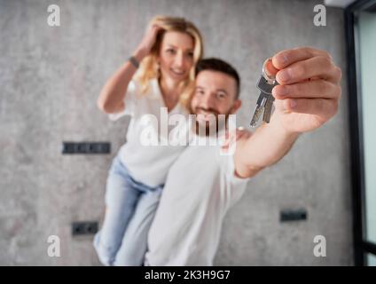 Ein fröhlicher Mann, der seine Frau mit einem Lächeln auf den Lippen hielt und dabei die Schlüssel zeigte. Glückliches Paar Immobilienkäufer demonstrieren Schlüssel von neuem Haus. Mit den Tasten auf die männliche Hand fokussieren. Stockfoto
