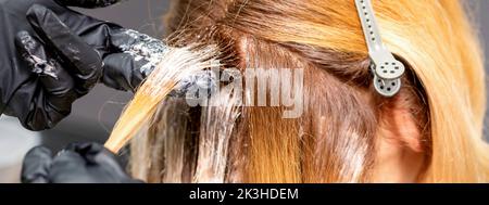 Die Hände des Friseurs tragen den Farbstoff mit Handschuhen auf das rote Haar einer jungen rothaarigen Frau in einem Friseursalon aus nächster Nähe auf Stockfoto