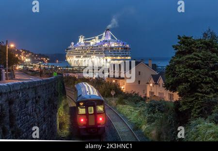 Cobh, Cork, Irland. 27.. September 2022. Ein Pendlerzug am frühen Morgen nähert sich dem Bahnhof, als das Kreuzschiff Norwegian Dawn an ihrem Liegeplatz in Cobh, Co. Cork, Irland, ankommt. - Bild David Creedon Stockfoto