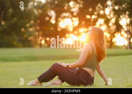 Junge Asiatin mit langen welligen Haaren, grünem Tank Top und schwarzer Leinenhose sitzt auf dem Gras und sonnt sich in der Sonne, die bei Sonnenuntergang durch die Bäume kommt. Stockfoto