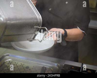 Koch in weißer Uniform gießt gekochte Spaghetti in den Topf-Wok, um Pasta mit Gemüse zu kochen Stockfoto