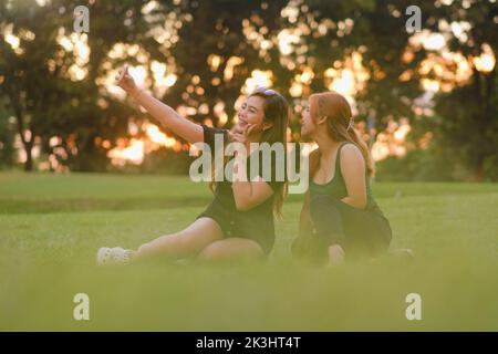 Zwei junge asiatische Frauen posieren für die Kamera, während sie zusammen ein Selfie machen. Beide sitzen an einem schönen Sommertag bei Sonnenuntergang auf dem Rasen in einem Park. Stockfoto