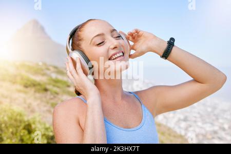 Fitness, Natur und eine Frau mit Kopfhörern, die Musik mit Lächeln und Berghintergrund hört. Übung, Streaming-App und eine Workout-Wiedergabeliste Stockfoto