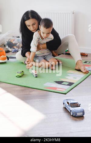 Behinderter Junge spielt Spielzeugautos mit Mutter zu Hause. Zerebrale Lähmung Kind unterhaltsam auf der Matte mit Betreuer. Kommunikation und Rehabilitation in Stockfoto