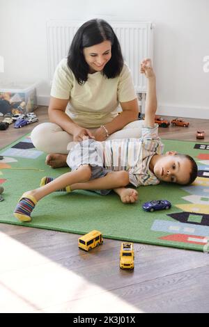 Behinderter Junge spielt Spielzeugautos mit Mutter zu Hause. Zerebrale Lähmung Kind unterhaltsam auf der Matte mit Betreuer. Kommunikation und Rehabilitation in Stockfoto