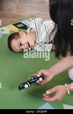 Behinderter Junge spielt Spielzeugautos mit Mutter zu Hause. Zerebrale Lähmung Kind unterhaltsam auf der Matte mit Betreuer. Kommunikation und Rehabilitation in Stockfoto