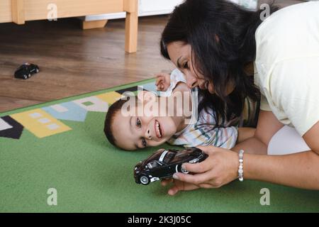 Behinderter Junge spielt Spielzeugautos mit Mutter zu Hause. Zerebrale Lähmung Kind unterhaltsam auf der Matte mit Betreuer. Kommunikation und Rehabilitation in Stockfoto