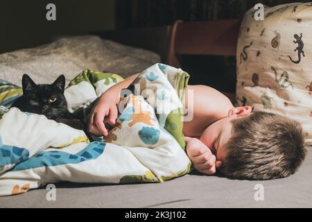 Kind, Junge schläft im Bett mit schwarzer Katze. Niedliches Kätzchen, das mit Kind auf dem Bett liegt und die Kamera anschaut. Zweisamkeit Kinder und Haustiere. Stockfoto