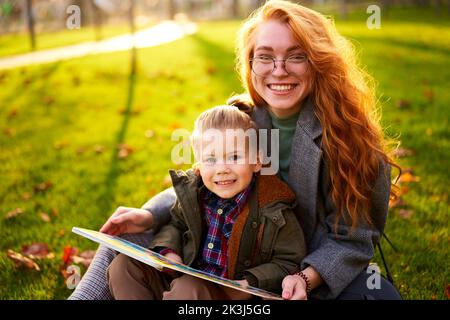 Rotschopf Frau liest Buch mit kleinen Jungen auf Gras im Stadtpark sitzen. Die junge Mutter unterrichtet ihren Sohn mit der ersten Klasse und macht Hausaufgaben am sonnigen Herbsttag Stockfoto