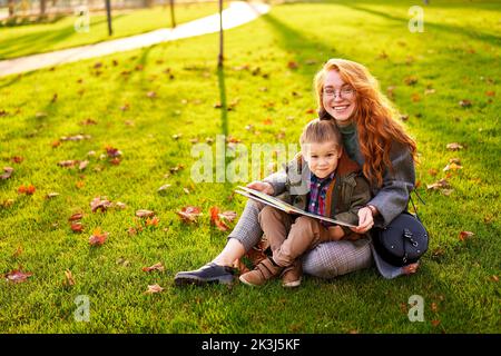 Rotschopf Frau liest Buch mit kleinen Jungen auf Gras im Stadtpark sitzen. Die junge Mutter unterrichtet ihren Sohn mit der ersten Klasse und macht Hausaufgaben am sonnigen Herbsttag Stockfoto