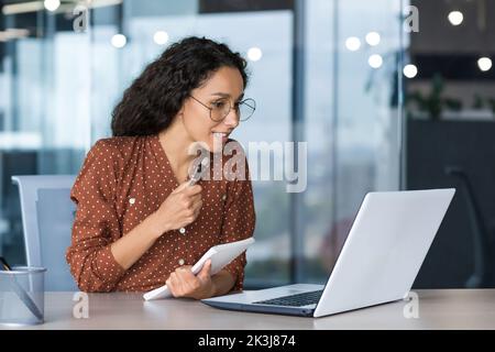 Junge, schöne und erfolgreiche Geschäftsfrau, die aus der Ferne im Büro sitzt, Hispanic-Frau, die mit einem Laptop für Videoanruf im Notebook schreibt, mit Kollegen und Mentoren aus der Ferne spricht Stockfoto