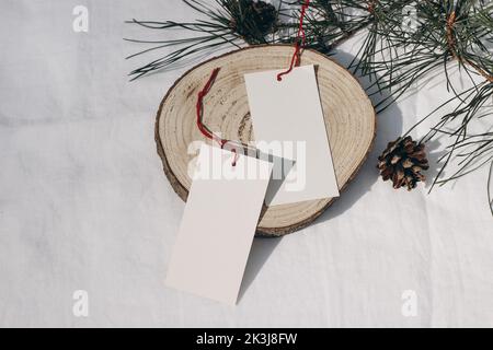 Weihnachtliche Sesonale Verkaufskonzept. Unbeschriftete Geschenkanhänger aus Papier, Etiketten mit rotem Seil auf geschnittenem Rundbrett aus Holz. Weiße Leinen Tischtuch Hintergrund mit Stockfoto