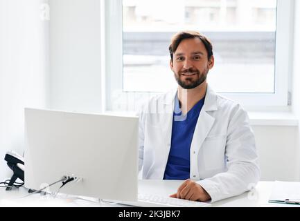 Porträt eines lächelnden Hausarztes in Arztuniform, der in seinem Arbeitszimmer in der medizinischen Klinik sitzt. Arztpraxis Stockfoto