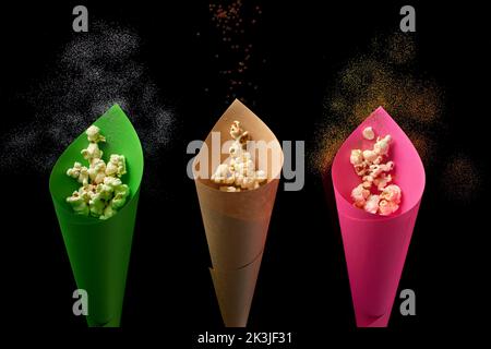 Grüne, braune und pinke Papierkegel voller Popcorn mit magischem Staub und Sternen auf schwarzem Hintergrund Stockfoto