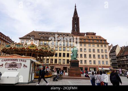 Menschen, die auf dem Gutenberg-Platz mit einer Statue von Johannes Gutenberg mit dem hinter Gebäuden ragenden Kirchturm spazieren, Straßburg, Frankreich. Stockfoto