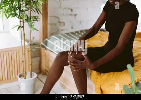 afroamerikanische Frau, die im dekorierten Studio sitzt und die Kniekappe mit den Händen massiert. Schwarze Person mit Sportverletzung, Beinmuskelbelastung. Schlechte Muskeln Stockfoto