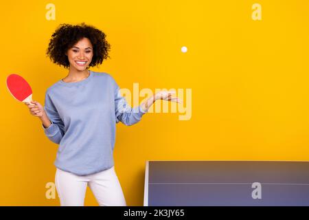 Foto von lustigen niedlichen lockigen Mädchen gekleidet blauen Pullover werfen Ball spielen Tischtennis isoliert gelben Hintergrund Stockfoto