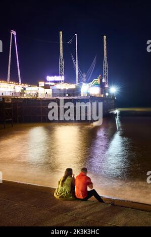 Während der Blackpool-Illuminationen saßen zwei Personen auf der Ufertreppe mit Blick auf den South Pier in der Nacht Stockfoto