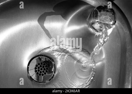 Glänzendes Waschbecken aus Stahl mit Wasser, das den Abfluss hinunterfließt Stockfoto
