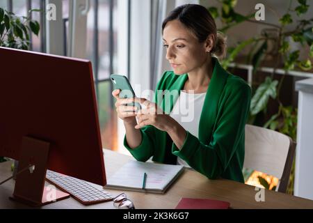 Nachdenkliche Geschäftsfrau sitzt am Tisch vor dem Computer, nutzt Smartphone für die Arbeit, scrollt Nachrichten. Stockfoto