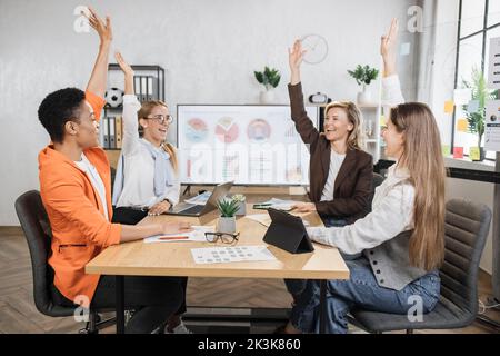 Ein Team aus multiethnischen Finanzexperten feiert erfolgreiche Verhandlungen, während sie am Schreibtisch sitzen und die Hände hoch halten. Frauen verwenden moderne Geräte für die Arbeit im Büro. Stockfoto