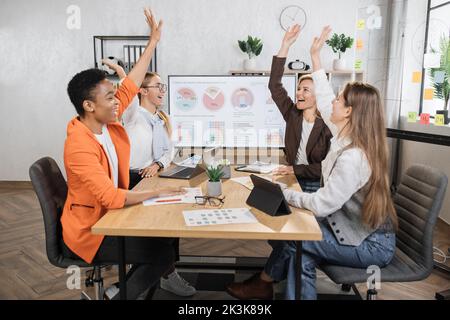 Ein Team aus multiethnischen Finanzexperten feiert erfolgreiche Verhandlungen, während sie am Schreibtisch sitzen und die Hände hoch halten. Frauen verwenden moderne Geräte für die Arbeit im Büro. Stockfoto