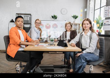 Weibliche Ökonomen in formeller Kleidung blicken auf die Kamera, während sie mit modernen Gadgets und Dokumenten zusammen am Schreibtisch sitzen. Multiethnische Maklerinnen verbringen den Abend, um den Handelsmarkt zu untersuchen. Stockfoto