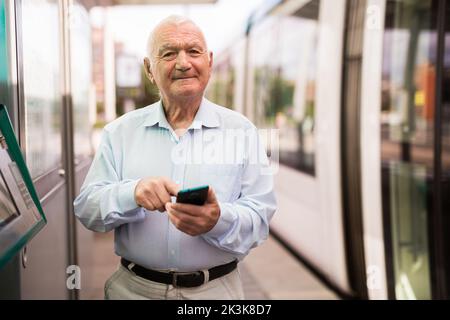 Alter Mann mit Smartphone an der Straßenbahnhaltestelle Stockfoto