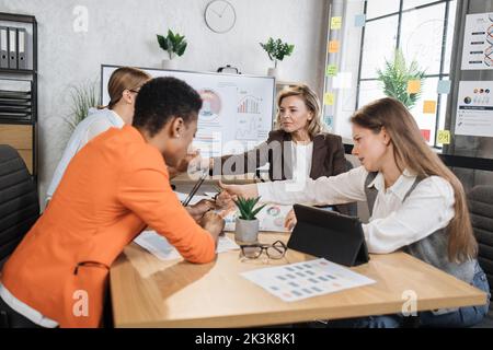 Vier multiethnische Ökonomen sitzen mit verschiedenen Gadgets im Büro und analysieren das Finanzwachstum in der weltweiten Wirtschaft. Weibliche Partner, die Geschäftstreffen abhalten. Stockfoto