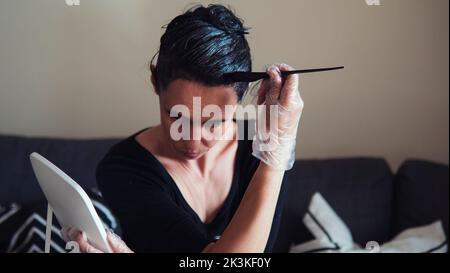 Frau Färbung dunkle Haare mit grauen Wurzeln zu Hause. Frau, die auf dem Sofa sitzt und mit einem schwarzen Pinsel Haare färben und auf die Spiegelglocke schaut Stockfoto