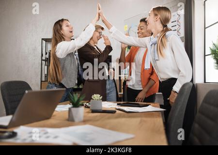 Verschiedene weibliche Finanztraderinnen geben sich nach produktiver Zusammenarbeit im Büro hohe fünf. Kompetente Makler, die neben dem Schreibtisch mit verschiedenen modernen Technologien und Papieren stehen. Stockfoto
