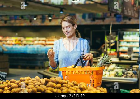 Junge schöne blonde Frau in einem blauen Pullover mit einem Korb mit Früchten in der Hand. Stehen an der Gemüseabteilung, Kartoffeln auswählen. Stockfoto