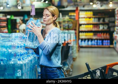 Eine junge Frau kauft in einem Supermarkt Wasserpaletten. Er nimmt es in die Hände, packt es in einen Wagen. Panik während einer Pandemie, Quarantäne. Stockfoto