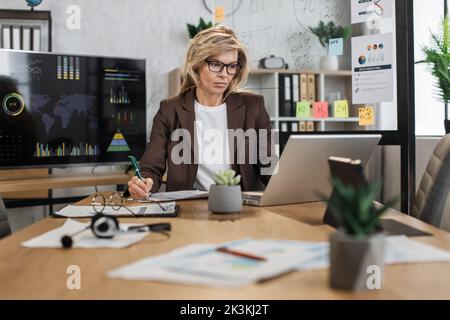 Porträt einer charmanten älteren Frau in elegantem Anzug, in dem Notizen geschrieben und ein moderner Laptop am Schreibtisch sitzt. Hübscher Firmenmitarbeiter mit blonden Haaren, die auf den Bildschirm schauen. Stockfoto