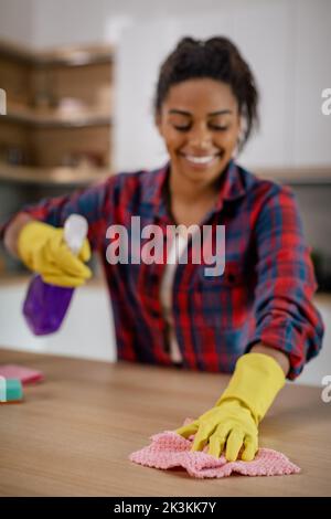 Fröhliche junge schwarze Dame wischt Tisch von Schmutz mit Reinigungsmitteln in der weißen Kücheneinrichtung, Nahaufnahme Stockfoto