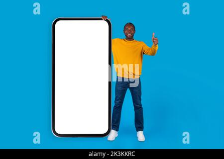 Fröhlicher schwarzer Mann, der neben einem großen Smartphone steht und den Daumen nach oben zeigt Stockfoto