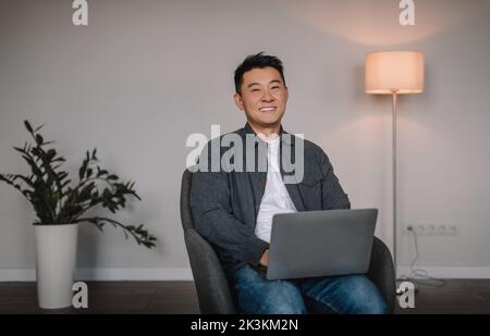 Fröhlicher chinesischer Mann mittleren Alters, der auf dem Laptop tippt, aus der Ferne online chattet, in einem Sessel sitzt Stockfoto