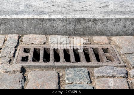 Gebrauchte Eisen rechteckige Kanalentwässerung auf der alten gepflasterten Straße in der Nähe der Granit-Bordstein und Steinfliesen Fußgänger Bürgersteig in der Nähe bei sonnigem Stockfoto