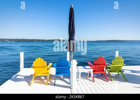 Vier farbige Holzstühle und ein blauer Sonnenschirm auf einem weißen Pier aus Holz, Genfersee in der Nähe von Fontana, Wisconsin, Amerika. Stockfoto