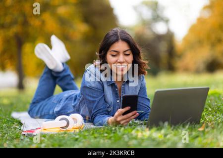 Lächelnde Junge Arabische Frau, Die Sich Mit Laptop Und Smartphone Im Park Entspannt Stockfoto