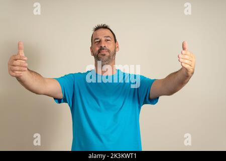 Kostenlose Umarmungen für die Person, die einen schlechten Tag hatte. Fröhliches, freundliches männliches Model in blauem T-Shirt, das die Hände zur Kamera zieht, sich umarmen und lächeln will Stockfoto