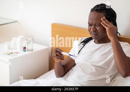 Sick Young African American Man Hält Thermometer, Während Im Bett Sitzend Stockfoto
