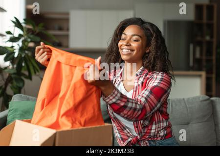 Glückliche hübsche junge afroamerikanische Frau öffnet Box, schauen Sie sich neue Kleidung an, genießt Kauf im Wohnzimmer Stockfoto