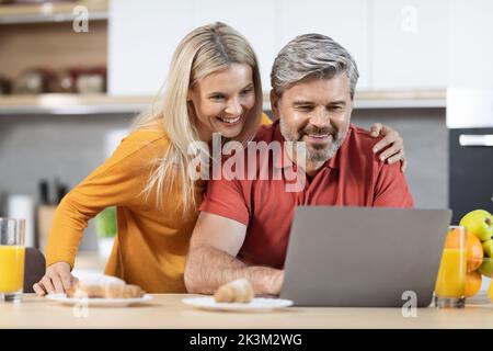 Glückliche Ehegatten, die einen Laptop benutzten, während sie in der Küche frühstückten Stockfoto